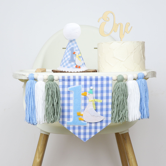 A complete 1st birthday high chair decoration setup. The chair is dressed with the blue gingham high chair banner and features a birthday cake with a wooden 'One' topper and a matching duck-themed party hat sitting on the tray.