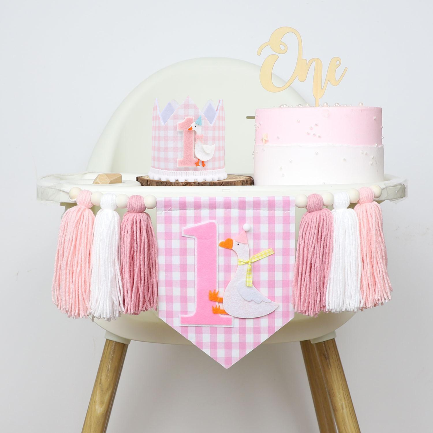 A complete 1st birthday high chair decoration setup. The chair is dressed with the pink gingham high chair banner and features a birthday cake with a wooden 'One' topper and a pink gingham party crown sitting on the tray.